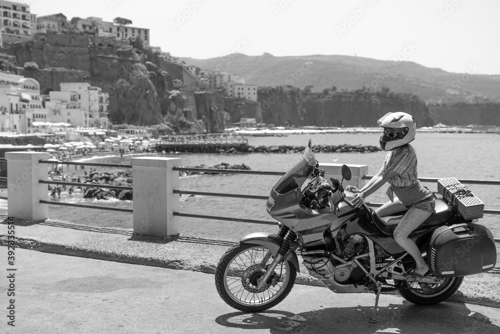 Young girl posing. Motorcycle for tourism and travel. Wearing a helmet. In the background are views of the sea and hotels. Coast of Italy. Sunny day, vacation and trip. Black and white