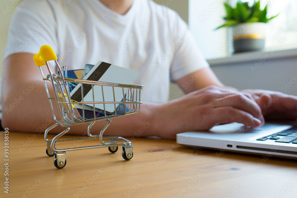Shopping cart for a supermarket with credit cards on the background of a man who work on a laptop. Online shopping concept