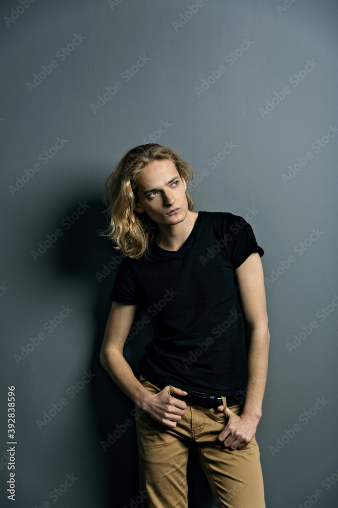 Young attractive guy with long blonde hair is sad leaning against a gray wall, looks to the camera Young attractive guy with long blonde hair is sad leaning against a gray wall, looks to the camera