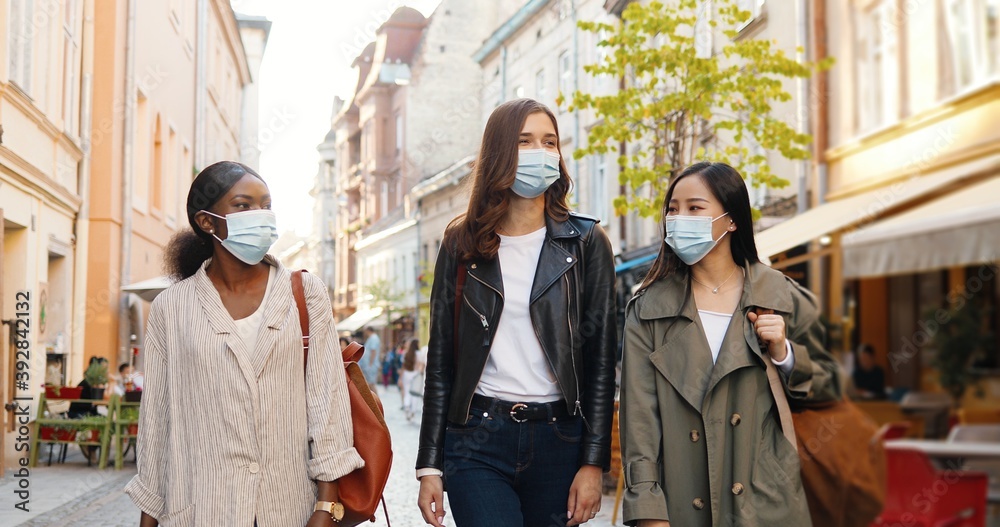 Obraz premium Three mixed-races young stylish women in medical masks talking and walking the street in city. Pretty cheerful multi ethnic females friends strolling outdoors and watching town. Tourists at pandemic.