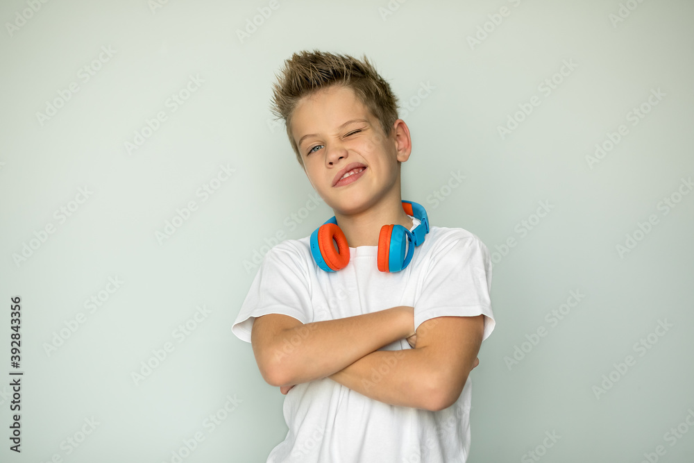 Handsome boy crossed his arms over his chest, one eye closed, funny. The teenager has headphones on his neck. The guy in the white shirt. Isolated. Handsome boy crossed his arms over his chest, one eye closed, funny. The teenager has headphones on his neck. The guy in the white shirt. Isolated.
