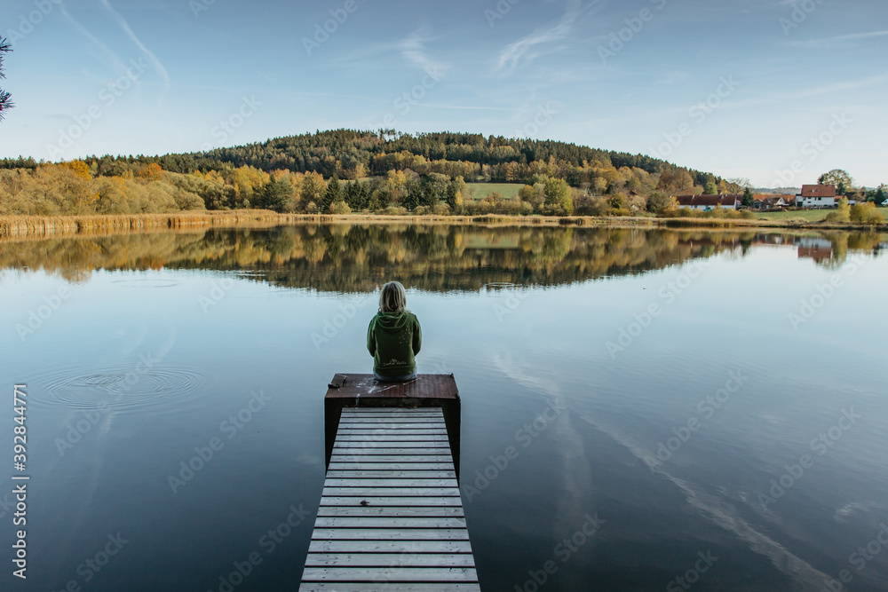 Calm morning relaxation by a pond. Sitting woman in the countryside. Intended female mysterious atmosphere. Spring sunny afternoon in the nature. Silence. Wooden pier on a pond. View of a fresh nature