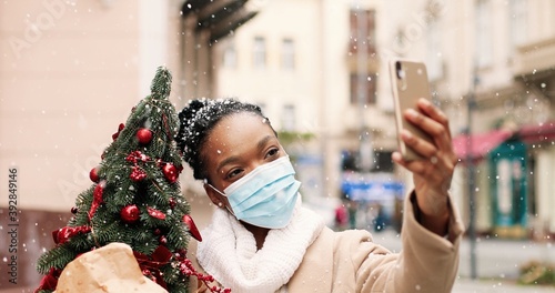 Close up portrait of African American happy female blogger in mask standing in snowy city and talking on video on smartphone. Woman with little new year tree outdoor videochatting on cellphone © VAKSMANV
