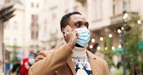 Close up portrait of happy young man in medical mask standing in snowy decorated city and calling on smartphone. African American male with christmas present talking on cellphone. X-mas spirit concept