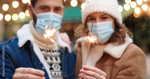 Close up of Caucasian happy couple holding sparklers while standing on decorated xmas street in medical masks in quarantine. Joyful man and woman celebrating new year 2021. Holiday celebration concept