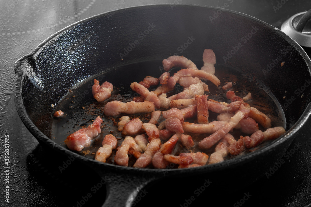 Chopped bacon lardons cooking on a hob in a cast iron frying pan Chopped bacon lardons cooking on a hob in a cast iron frying pan