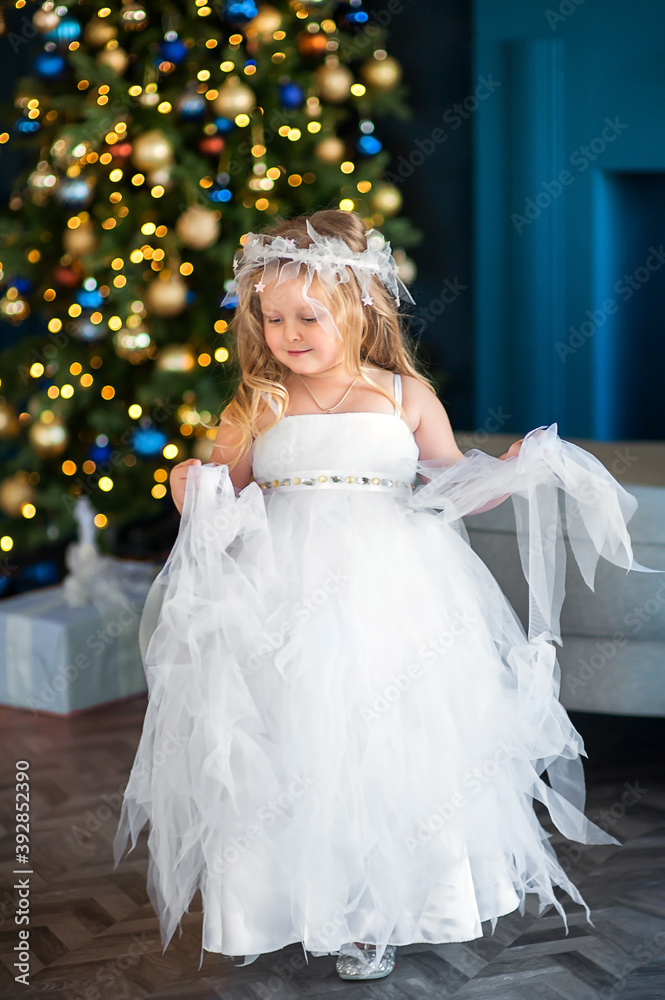 Cute little girl blonde with blue eyes, looking like an angel in a white dress, near the Christmas tree Cute little girl blonde with blue eyes, looking like an angel in a white dress, near the Christmas tree