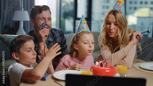 Girl blowing candles on cake with parents. Family celebrating birthday online.