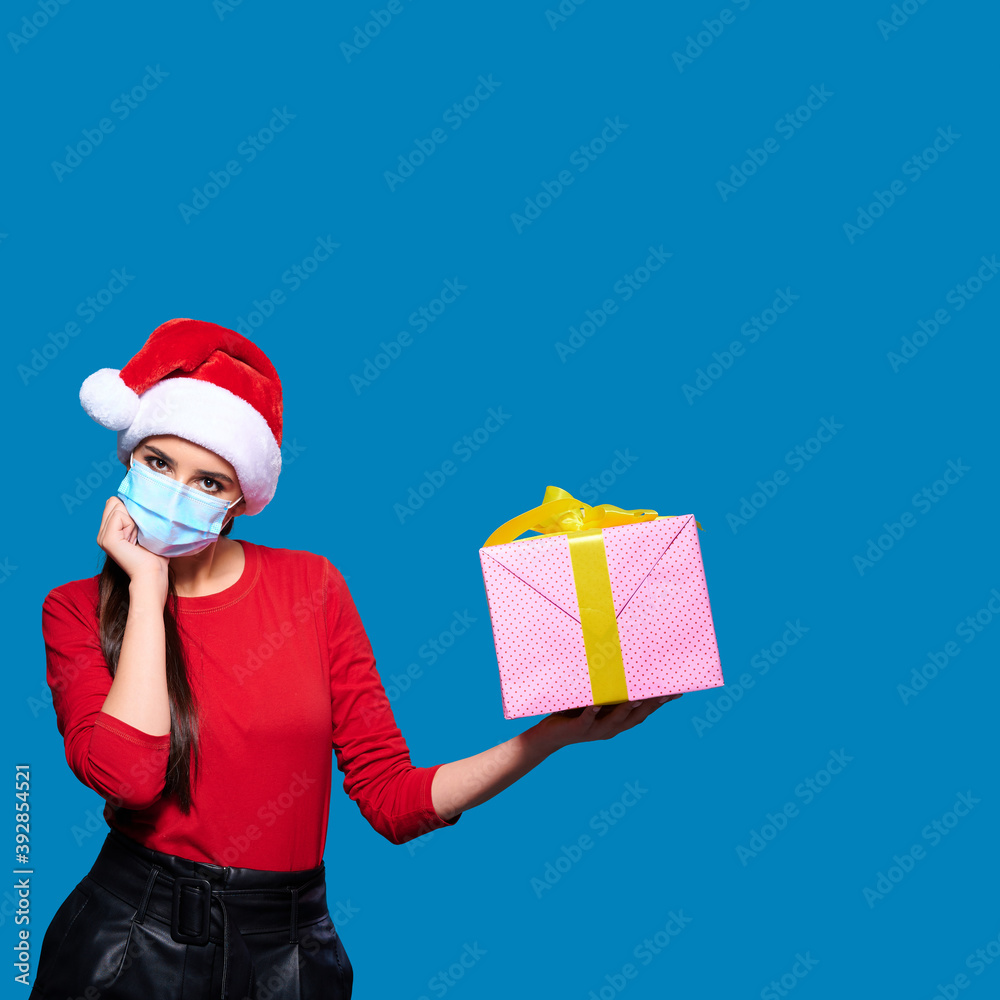 Upset woman wearing Santa hat and face protective medical mask and touching face, blue isolated background. Upset woman wearing Santa hat and face protective medical mask and touching face, blue isolated background.
