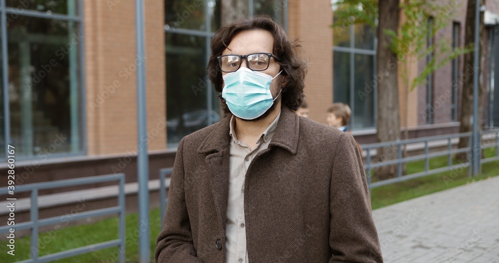 Portrait of serious Caucasian handsome man educator in glasses an mask standing in front of school with pupils on background. Male teacher with planner outdoors in quarantine. Education concept Portrait of serious Caucasian handsome man educator in glasses an mask standing in front of school with pupils on background. Male teacher with planner outdoors in quarantine. Education concept