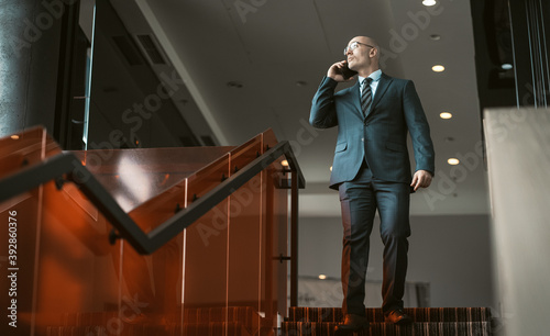 Businessman talking on cell phone while standing at top of stairs in business center. Handsome Caucasian man looking at copy space on left side. Career growth concept.