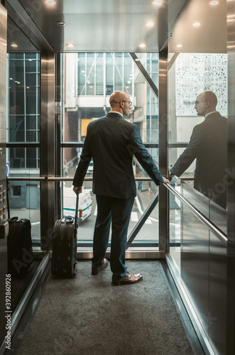 Businessman with suitcase in business center. Business trip concept. Middle aged Caucasian man looks out through glass wall of elevator. Rear view.