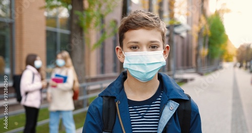 Close up portrait of joyful cute schoolboy standing outdoors with backpack and putting off medical mask. Male Caucasian student in mask near school smiling to camera. Quarantine concept
