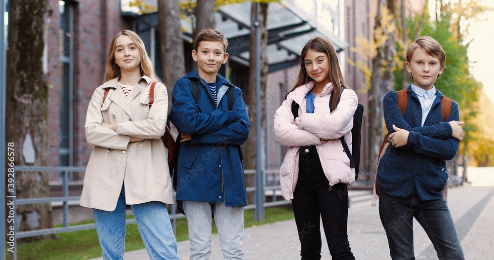 Caucasian cheerful school kids standing on street near school and posing to camera while smiling in good mood. Happy girls and boys pupils with backpacks outdoor. Classmates concept