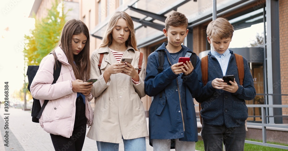 Caucasian concentrated teens standing outdoors and typing on cellphones. Beautiful girl pupil browsing on smartphone near school. Young boy school student texting on mobile phone. Friends concept