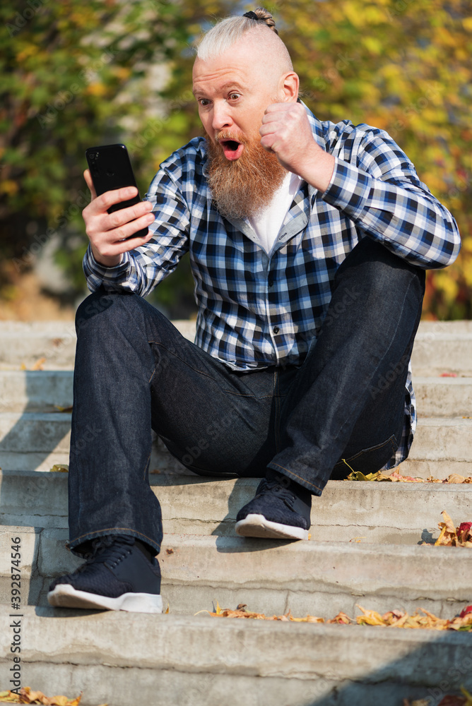 Excited overjoyed bearded man looking at smartphone screen sitting on steps outdoors feeling euphoric celebrating victory. Winner concept.