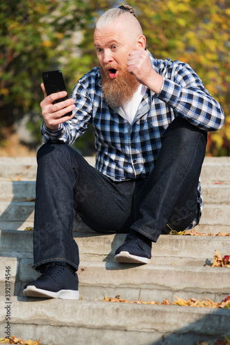 Obraz na płótnie Excited overjoyed bearded man looking at smartphone screen sitting on steps outdoors feeling euphoric celebrating victory. Winner concept. w salonie
