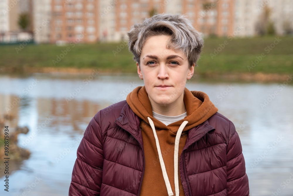 Young woman with stylish short hair, dressed in a gender neutral style. Portrait of a woman against the background of a residential building and a pond Young woman with stylish short hair, dressed in a gender neutral style. Portrait of a woman against the background of a residential building and a pond