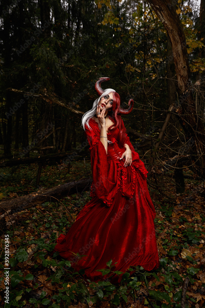 Maleficent Woman in Red Clothing and Horns in dark Forest. Posing in magik forest