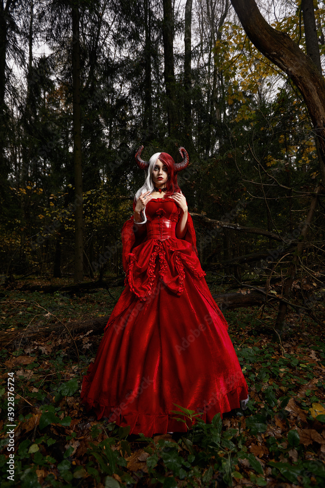 Maleficent Woman in Red Clothing and Horns in dark Forest. Posing in magik forest