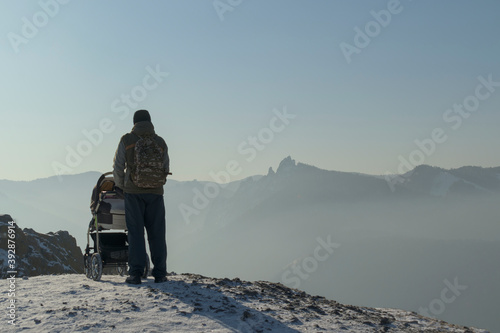 Man with a baby carriage stands on the top in the mountains. View from the back. Winter, white snow. Bright sun, shadows. The background is the sky. The concept of fatherhood, winter tourism.