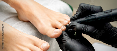 Pedicure master removes cuticle from toes of woman using professional electric nail hardware in nail salon