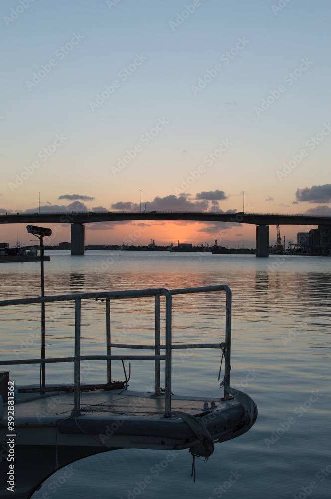 Fototapeta premium オレンジ色の夕焼けとシルエットの高架橋のある凪の港と船の先端