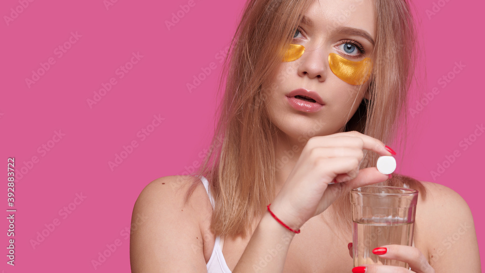 Woman with flowing hair puts pain reliever pill into glass of water after hangover on pink isolated background