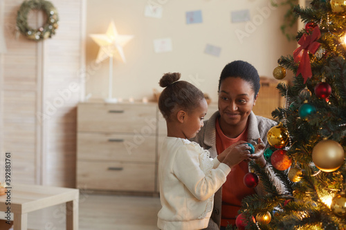 Side view portrait of cute African-American girl decorating Christmas tree with smiling happy mom in cozy home interior, copy space photo