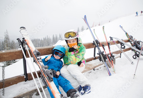 toddler boy and teenage girl ,brother and sister, are resting on bench, taking break from skiing in mountains. Winter sports for family. Children's skiing lesson at alpine school. Seasonal joys