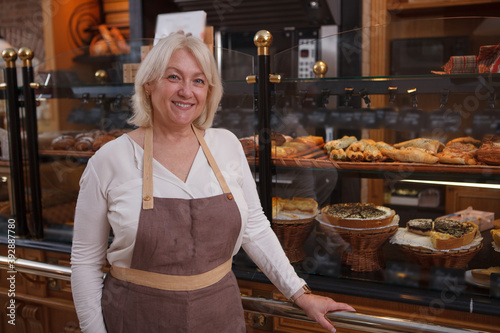 Charming mature female baker smiling to the camera, posing confidently at her bakery store, copy space