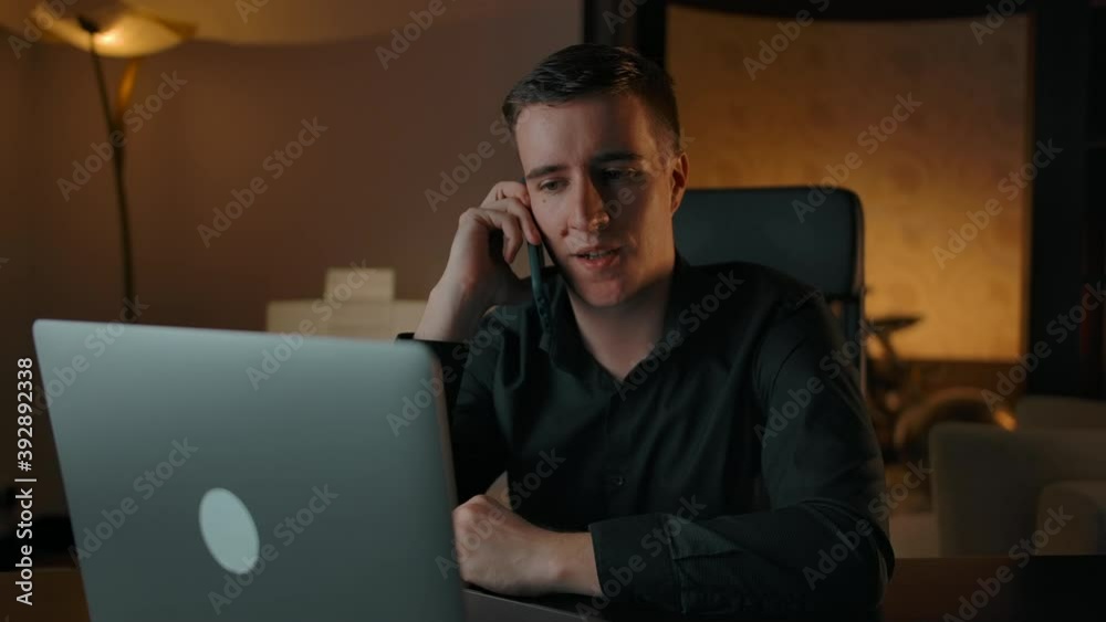 Caucasian man communicates on the phone sitting at his laptop at home. Cozy home interior. Finishing call and looking at computer display. Caucasian man communicates on the phone sitting at his laptop at home. Cozy home interior. Finishing call and looking at computer display.