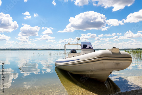Big white modern fishing motorboat moored at lake or river shore sand pebble beach against scenic blue sky on bright sunny summer day. Inflatable boat parked on pond bank after water surface trip