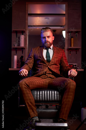 handsome young bearded man in a tuxedo sitting in dark room in fashionable clothing for the festive evening