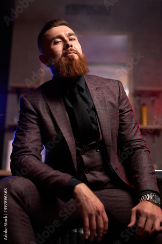 young caucasian handsome bearded man in suit sitting on couch in a dark smoky room, he is elegant and attractive, male is lost in thought. dramatic evening, dark shadows and wealth