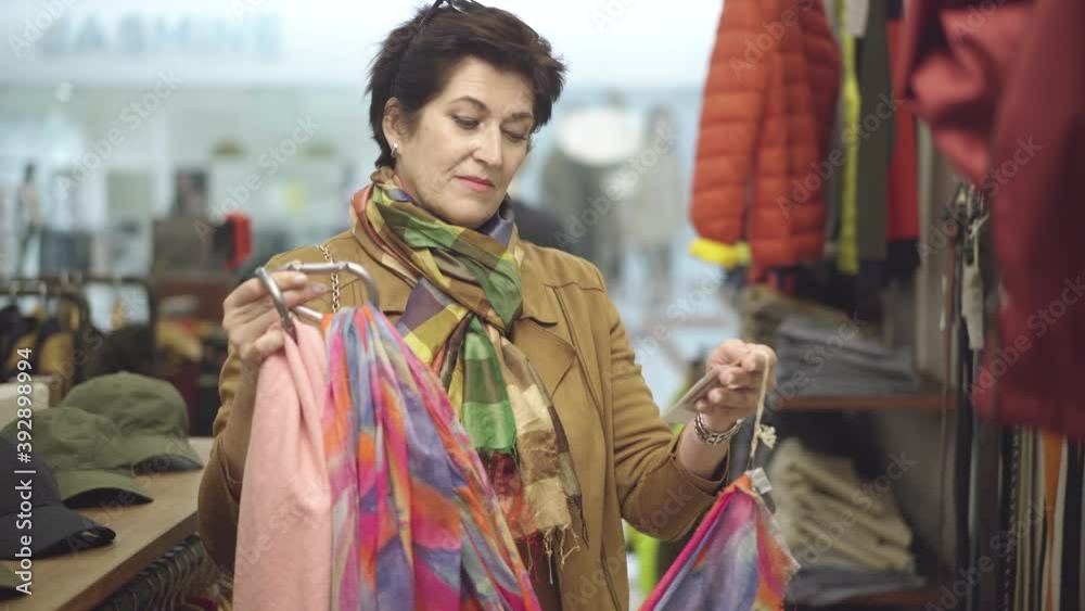 Portrait of elegant mid-adult female shopper choosing scarfs in clothing store. Confident Caucasian woman buying fashionable outfit during Black Friday sales. Lifestyle and consumerism concept. Portrait of elegant mid-adult female shopper choosing scarfs in clothing store. Confident Caucasian woman buying fashionable outfit during Black Friday sales. Lifestyle and consumerism concept.