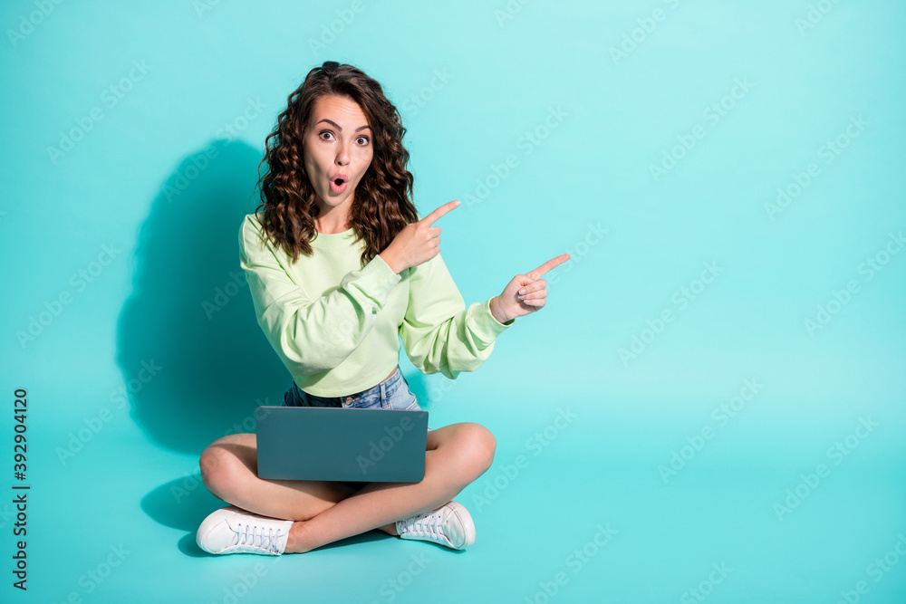 Photo of excited girl sit floor hold pc open mouth direct fingers empty space wear green sweatshirt sneakers isolated blue color background Photo of excited girl sit floor hold pc open mouth direct fingers empty space wear green sweatshirt sneakers isolated blue color background