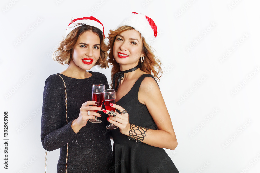 Two lovely  sexy girls  in red christmas santa claus holiday hat posing on white background, holding glass of wine.