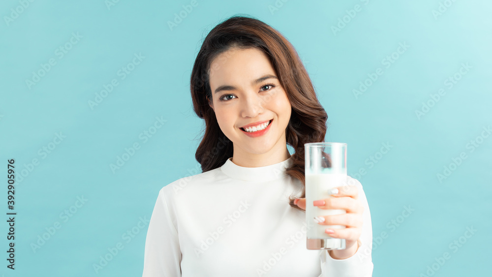 Young beautiful girl holding glass with milk in hand happily looking in camera over blue background Young beautiful girl holding glass with milk in hand happily looking in camera over blue background