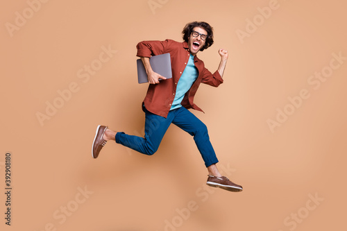 Photo portrait full body view of screaming guy in casual brown shirt running with laptop in arm jumping up isolated on pastel beige colored background