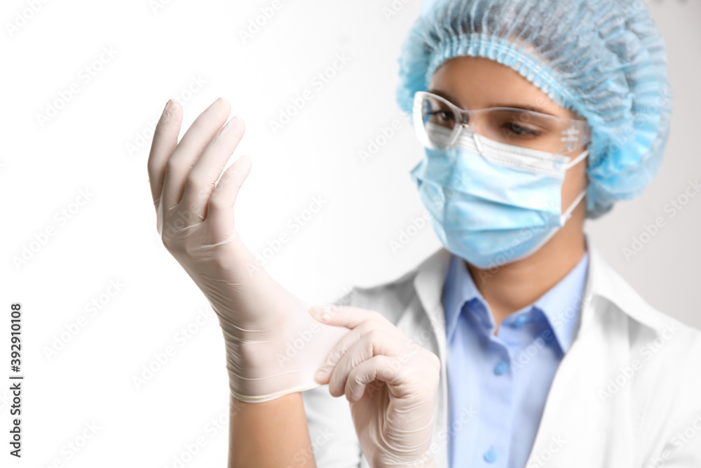 Woman in protective mask and glasses putting on medical glove against white background