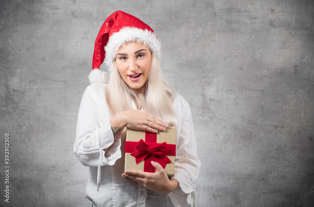 Fototapeta premium Happy beautiful blond woman wears christmas santa claus hat - cap and holding red gift box - present with ribbon, isolated on gray background