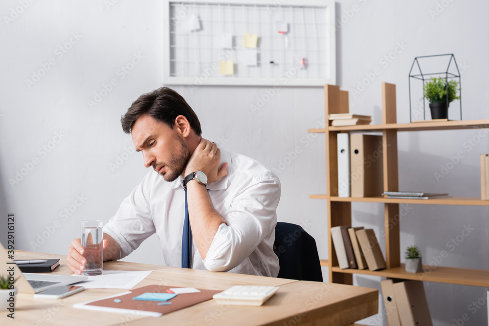 Businessman with painful neck holding glass of water, while sitting at workplace in office Businessman with painful neck holding glass of water, while sitting at workplace in office