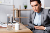 Glass of water and medication on desk with blurred businesswoman with stomachache on background
