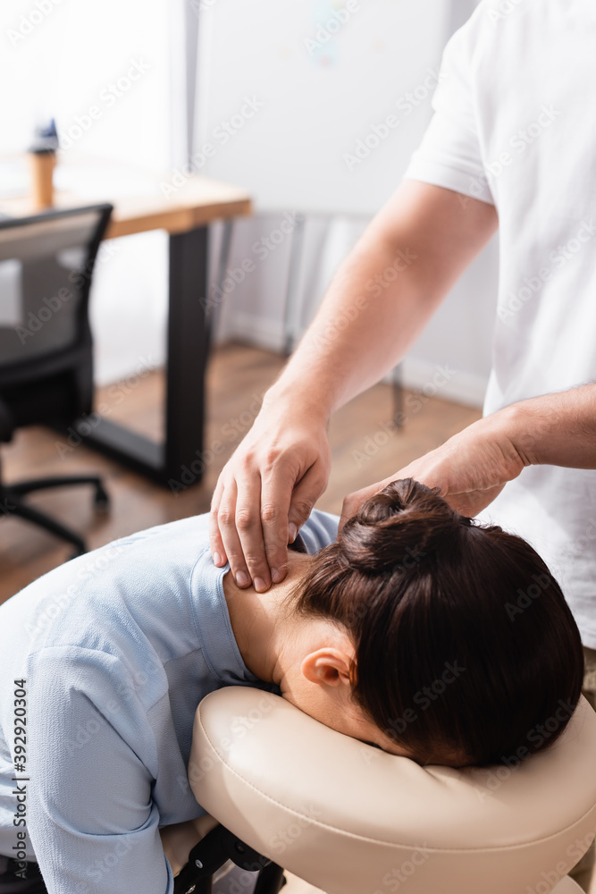 Massage therapist massaging neck of brunette businesswoman sitting on massage chair on blurred background Massage therapist massaging neck of brunette businesswoman sitting on massage chair on blurred background