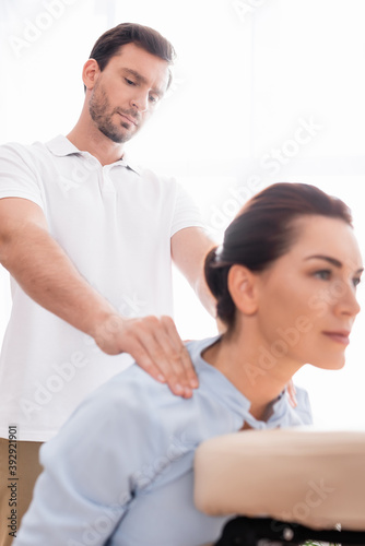 Focused masseur doing shoulders massage of positive female client on blurred foreground