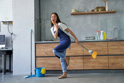 Happy worker. Full length shot of cheerful young woman, cleaning lady pretending to sing a song, holding broom while cleaning the floor, doing household chores photo