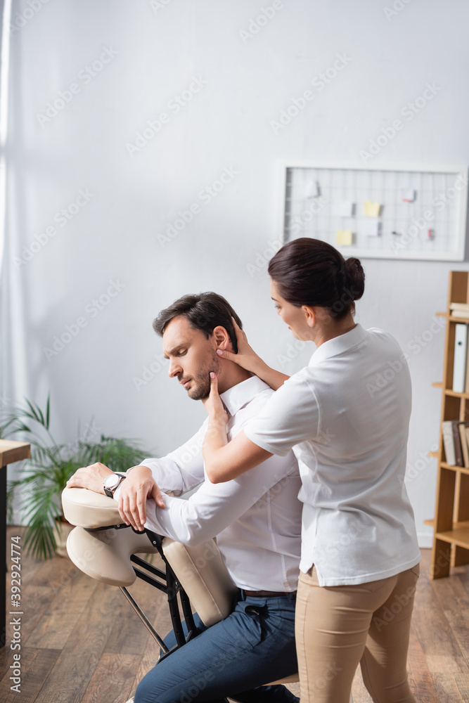 Masseuse massaging head of businessman sitting on massage chair in office on blurred background Masseuse massaging head of businessman sitting on massage chair in office on blurred background