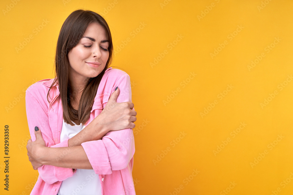 Obraz premium Love and hug yourself concept. Brunette girl in shirt embraces herself, enjoys with closed eyes her new comfortable fragrant clothes isolated in pink studio