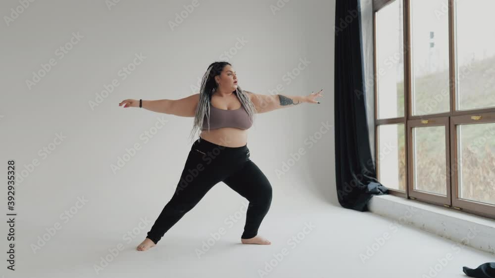 Happy body positive fat woman with dreadlocks doing yoga in the gym. Concept of natural diverse beauty and sport. Stout over weight female stretching isolated. Relaxation and tranquilty concept.